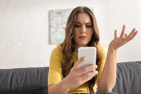 Atractiva mujer confundida usando teléfono inteligente en el sofá en la sala de estar - foto de stock