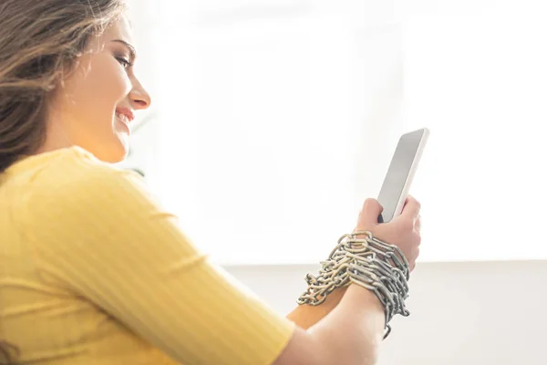 Side view of smiling woman holding smartphone in tied hands with metal chain — Stock Photo