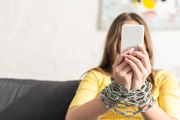 Selective focus of depended woman with tied hands with metal chain holding smartphone on couch — Stock Photo