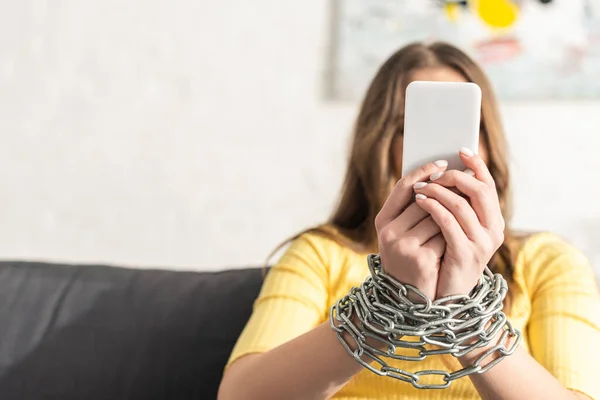 Selektiver Fokus einer abhängigen Frau mit gefesselten Händen mit Metallkette, die ihr Smartphone auf der Couch hält — Stockfoto