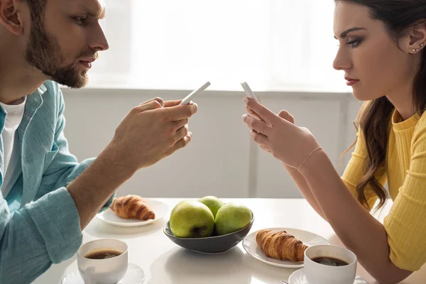 Vista lateral de la pareja utilizando teléfonos inteligentes cerca de café y cruasanes en la mesa - foto de stock