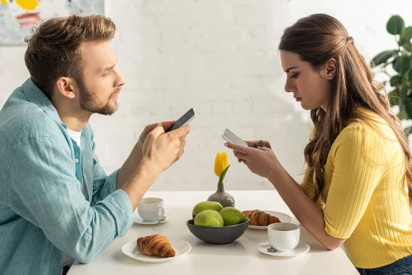 Vista lateral del hombre charlando cerca de novia celebración de teléfono inteligente durante el desayuno en la cocina - foto de stock