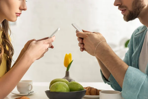 Vista recortada de pareja charlando con teléfonos inteligentes durante el desayuno en la cocina - foto de stock