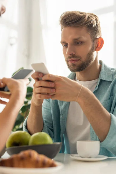 Foco seletivo de casal viciado segurando smartphones perto de café e croissant na mesa — Fotografia de Stock