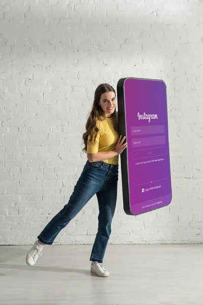 KYIV, UCRANIA - 21 de febrero de 2020: Mujer atractiva sonriendo a la cámara mientras sostiene el modelo de teléfono inteligente con la aplicación instagram - foto de stock