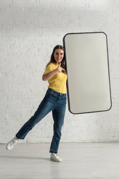 Chica sonriente apuntando con el dedo al modelo de teléfono inteligente cerca de la pared de ladrillo blanco - foto de stock