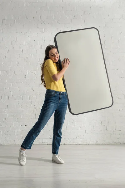 Femme gaie tenant le modèle de smartphone près du mur de briques blanches — Photo de stock