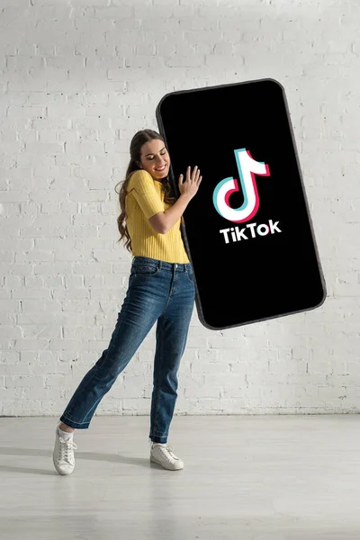 KYIV, UKRAINE - FEVEREIRO 21, 2020: Mulher feliz segurando modelo de smartphone com aplicativo TikTok perto da parede de tijolo branco — Fotografia de Stock