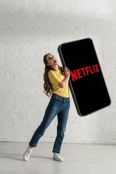 KYIV, UKRAINE - FEBRUARY 21, 2020: Attractive woman laughing while holding model of smartphone with netflix app at home — Stock Photo