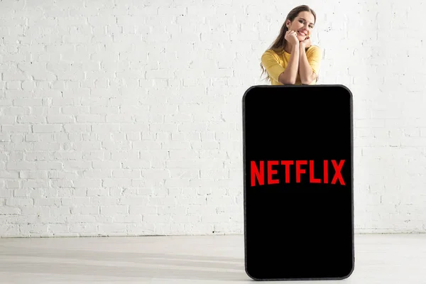 KYIV, UCRANIA - 21 de febrero de 2020: Muchacha sonriente mirando la cámara cerca de un gran modelo de teléfono inteligente con aplicación de netflix en casa - foto de stock