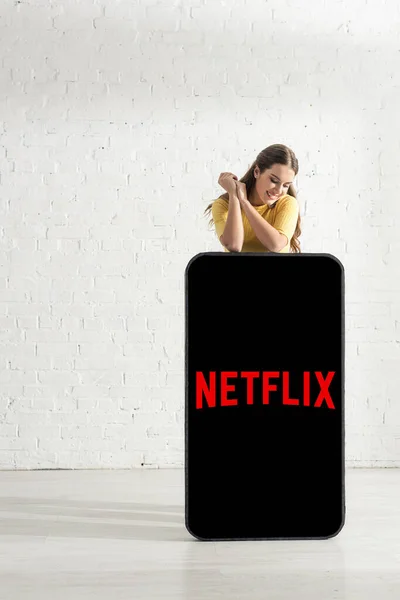 KYIV, UCRANIA - 21 de febrero de 2020: Muchacha atractiva sonriendo cerca del modelo de teléfono inteligente con la aplicación netflix - foto de stock