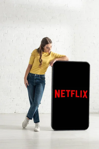 KYIV, UCRAINA - 21 FEBBRAIO 2020: Attraente giovane donna in piedi vicino al modello di smartphone con l'app netflix — Foto stock