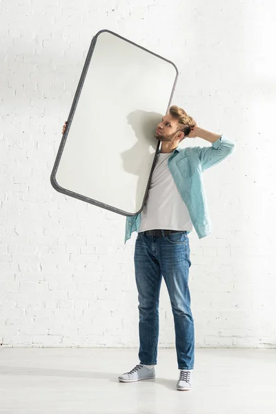Hombre pensativo con la mano cerca de la cabeza sosteniendo el modelo de teléfono inteligente en casa - foto de stock
