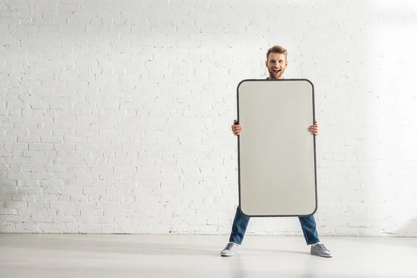 Homem alegre segurando grande modelo de smartphone perto da parede de tijolo branco — Fotografia de Stock