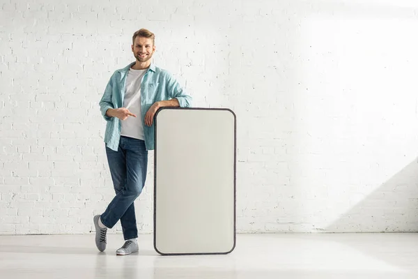 Hombre guapo sonriendo a la cámara y señalando con el dedo al gran modelo de teléfono inteligente en casa - foto de stock