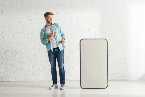 Испуганный мужчина смотрит на большую модель смартфона рядом с белой кирпичной стеной — стоковое фото