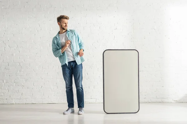 Hombre asustado mirando gran modelo de teléfono inteligente cerca de la pared de ladrillo blanco - foto de stock