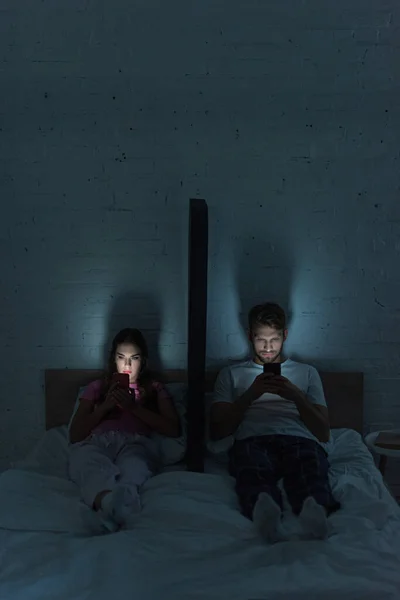 Young couple with smartphones sitting near model of smartphone on bed at night — Stock Photo
