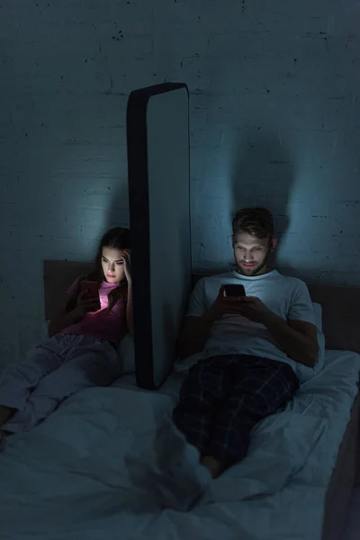 Couple using smartphones near model of smartphone on bed at night — Stock Photo