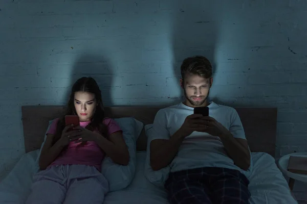 Young addicted couple using smartphones on bed at night — Stock Photo
