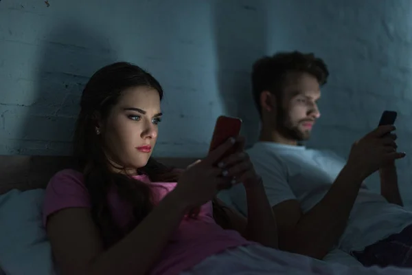 Selective focus of woman using smartphone near boyfriend chatting on bed at night — Stock Photo
