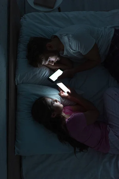 Вид зверху на молоду залежну пару, використовуючи смартфони, лежачи на ліжку вночі — стокове фото