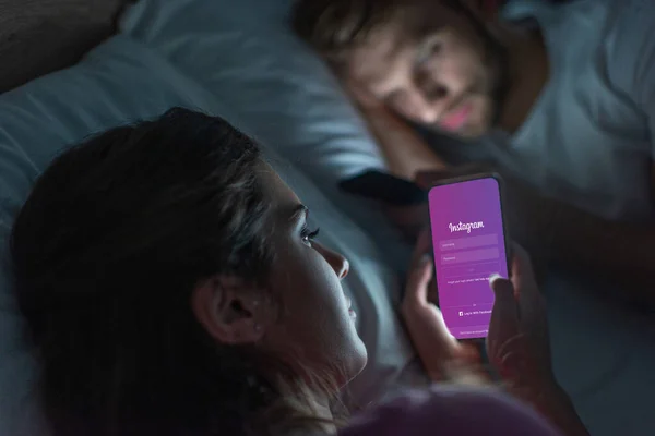 KYIV, UCRANIA - 21 de FEBRERO de 2020: Enfoque selectivo de la niña usando el teléfono inteligente con la aplicación instagram cerca del novio en la cama por la noche - foto de stock
