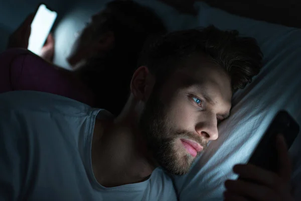 Selective focus of handsome man using smartphone near girlfriend on bed at night — Stock Photo