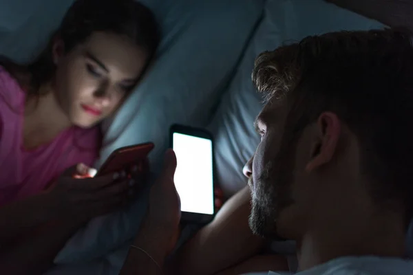 Selective focus of man using smartphone near girlfriend chatting on bed at night — Stock Photo