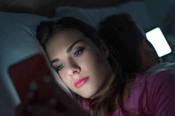 Selective focus of addicted woman using smartphone near man on bed at night — Stock Photo