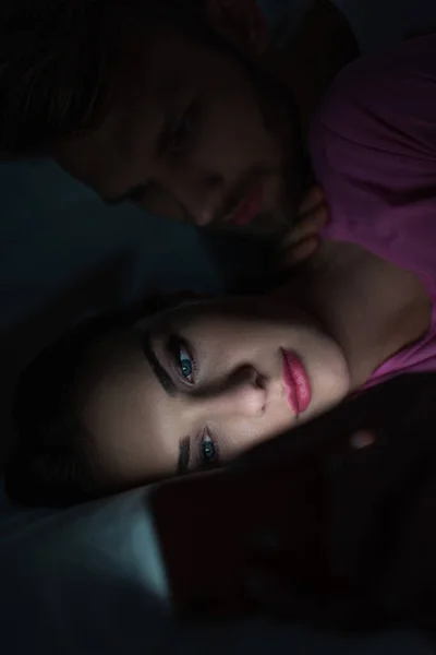 Selective focus of man embracing smartphone dependent woman on bed at night — Stock Photo