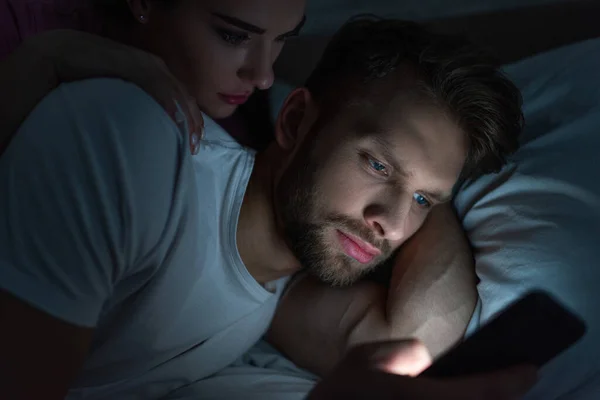 Enfoque selectivo de la mujer abrazando novio dependiente de teléfonos inteligentes en la cama por la noche - foto de stock