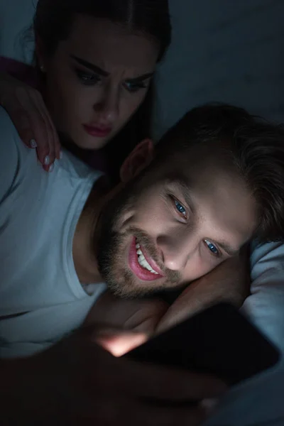 Mujer enojada abrazando novio sonriente con teléfono inteligente en la cama por la noche — Stock Photo