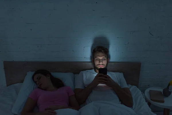 Smartphone-abhängiger Mann sitzt neben schlafender Freundin im Bett — Stockfoto