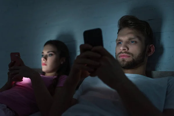 Selective focus of smartphone depended couple lying on bed at night — Stock Photo