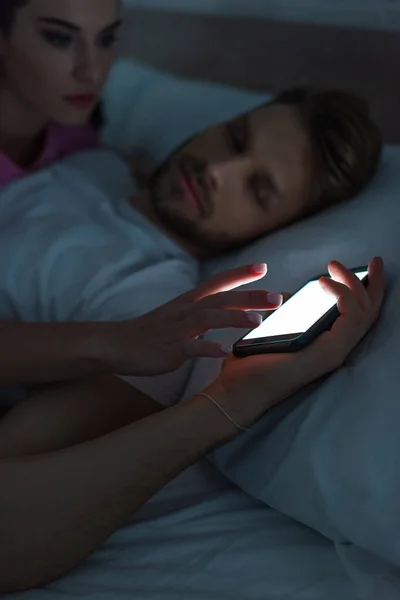 Selective focus of woman taking smartphone from sleeping boyfriend on bed at night — Stock Photo