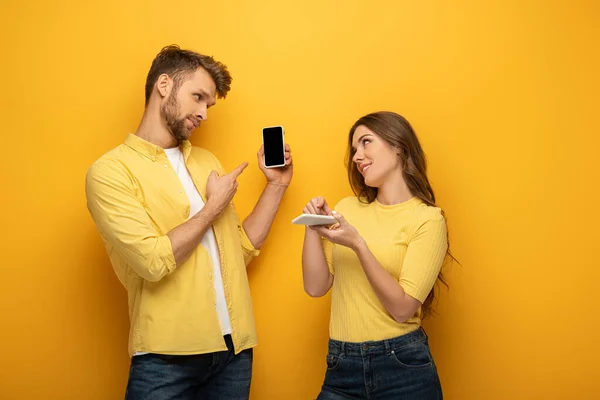 Giovane coppia che punta su smartphone mentre si guarda l'un l'altro su sfondo giallo — Foto stock