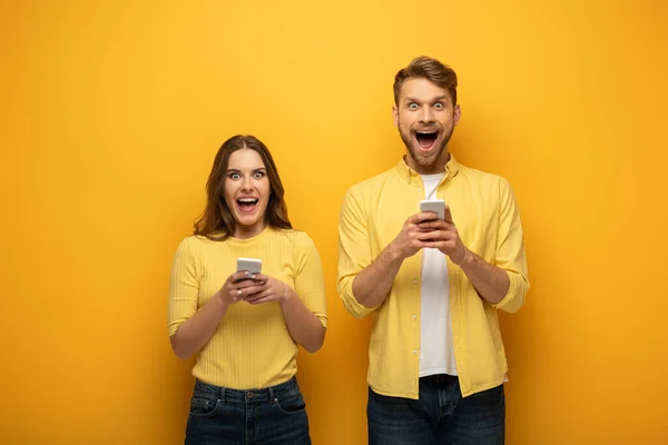 Aufgeregtes Paar blickt auf Kamera, während es Smartphones auf gelbem Hintergrund benutzt — Stockfoto