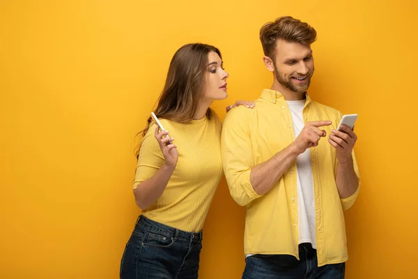 Sorrindo homem apontando com o dedo para smartphone perto da namorada no fundo amarelo — Fotografia de Stock