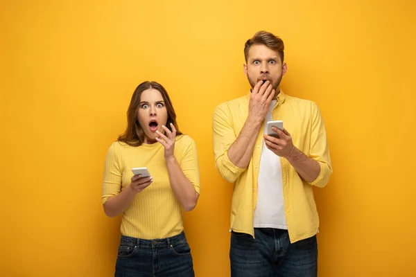 Shocked couple looking at camera while holding smartphones on yellow background — Stock Photo