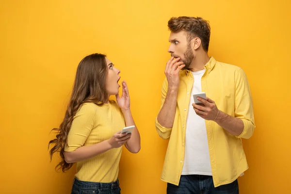 Surprised couple with smartphones looking at each other on yellow background — Stock Photo