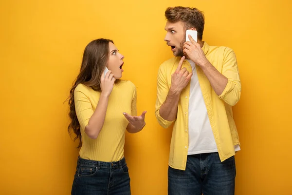 Shocked couple looking at each other while talking on smartphones on yellow background — Stock Photo