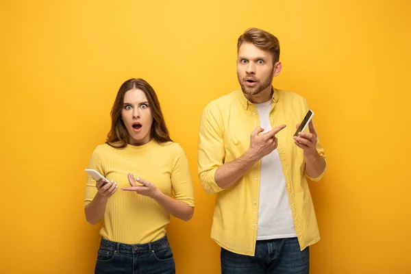 Pareja emocionada mirando a la cámara y apuntando a teléfonos inteligentes sobre fondo amarillo - foto de stock