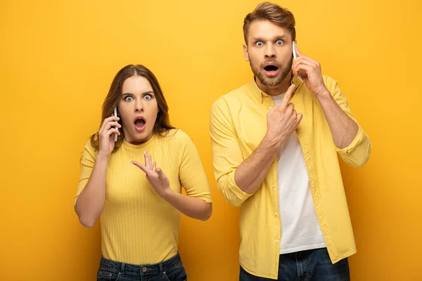 Überraschtes Paar zeigt mit dem Finger, während es auf Smartphones auf gelbem Hintergrund spricht — Stockfoto