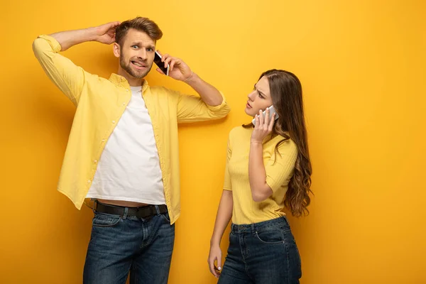 Skeptische Frau schaut verwirrten Mann an, der auf gelbem Hintergrund auf Smartphone spricht — Stockfoto