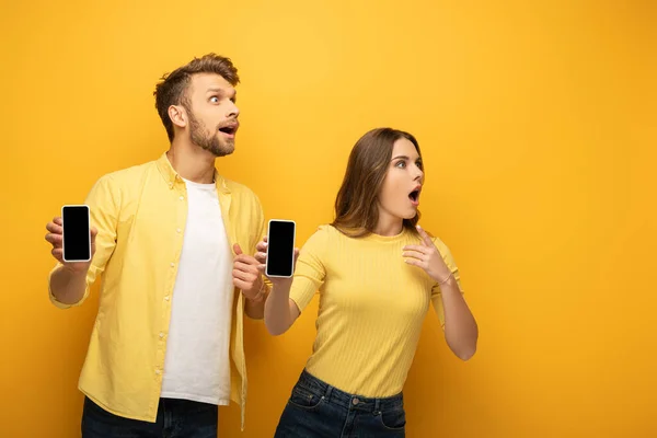 Shocked young couple looking away while showing smartphones with blank screens on yellow background — Stock Photo