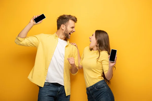 Emocionada pareja joven sosteniendo teléfonos inteligentes con pantallas en blanco sobre fondo amarillo - foto de stock