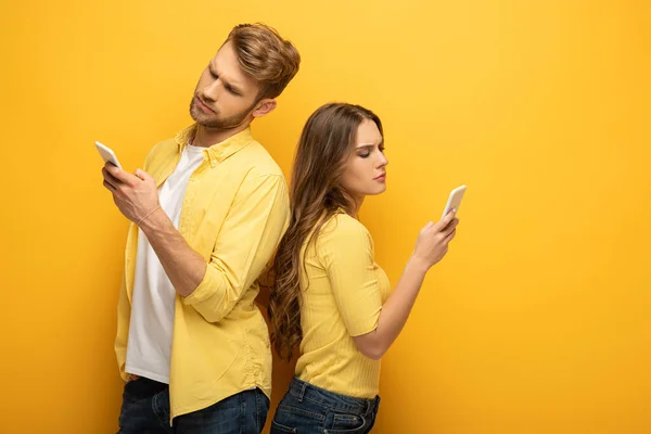 Casal jovem usando smartphones enquanto está de costas para trás no fundo amarelo — Fotografia de Stock