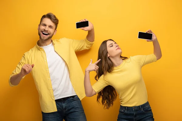 Positives Paar zeigt Smartphones mit leeren Bildschirmen auf gelbem Hintergrund — Stockfoto