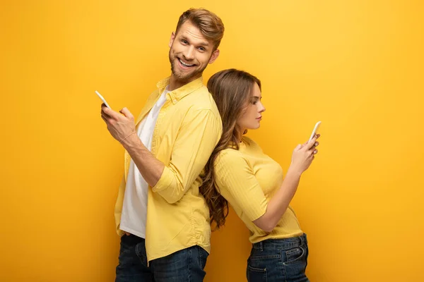 Vue latérale de l'homme souriant à la caméra près de petite amie en utilisant un smartphone sur fond jaune — Photo de stock